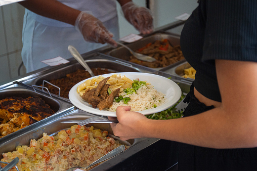 Imagem: Em primeiro plano e vestida de preto, aluna segura prato com refeição, em frente à bancada de serviço do Restaurante Universitário do Campus de Itapajé; ao fundo, um funcionário de branco a serve. (Foto: Viktor Braga/ UFC Informa)