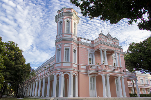 Foto da fachada da reitoria. É um prédio de cor rosa com detalhes brancos nas colunas e janelas. Ao fundo um céu azul cheio de pequenas nuvens. Ao redor da reitoria o verde das mangueiras. Fim da descrição.