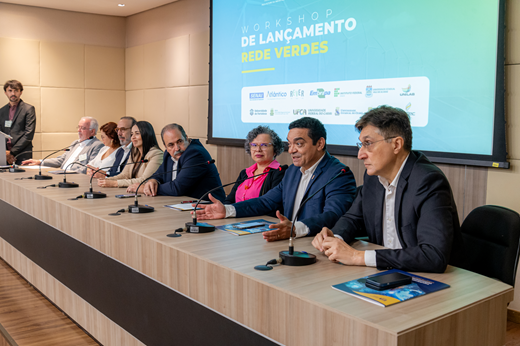 Imagem: Foto mostra o cerimonialista do evento em pé, no púlpito, e a mesa solene formada por oito pessoas sentadas. Ao fundo, um telão projeta apresentação com fundo verde. (Foto: George Lucas/ FIEC)