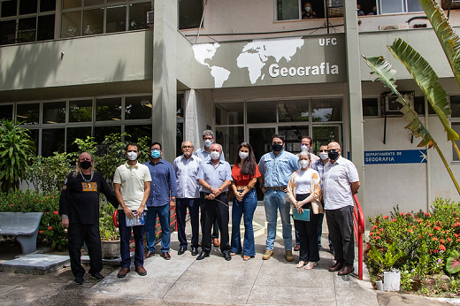 Imagem mostra grupo de pessoas em pé, lado a lado, posando para foto com prédio ao fundo com a identificação "Departamento de Geografia"