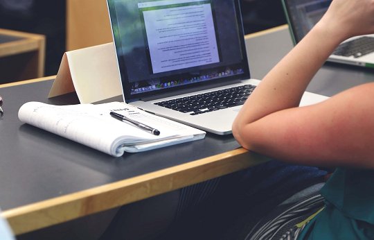 Imagem: Braço de uma pessoa apoiado sobre uma bancada, com um notebook e um caderno à frente (Foto: Pixnio)