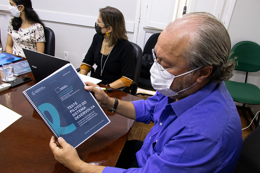 Imagem: foto de um homem branco de camisa azul segurando um material impresso encadernado com a capa azul marinho