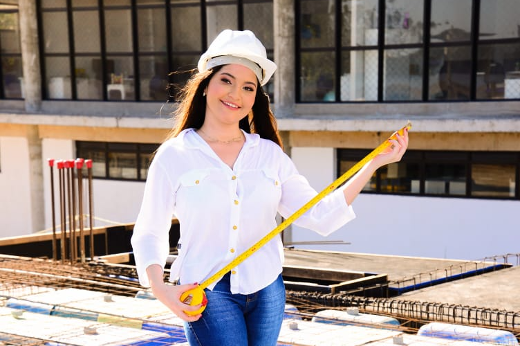 Imagem: Foto da engenheira civil Tereza Melo em uma obra, vestindo uma blusa branca, capacete branco e uma calça jeans azul e segurando uma trena amarela