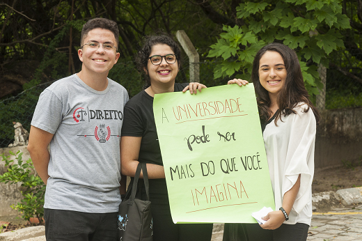 Imagem: Três estudantes, sendo que duas delas seguram um cartaz no qual está escrito "A universidade pode ser mais do que você imagina" (Foto: Viktor Braga/UFC) "