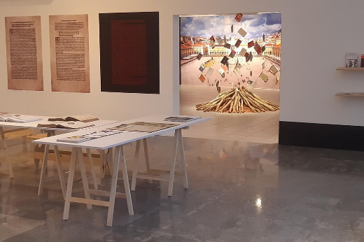 Imagem: Foto do interior da exposição com quadros, mesa com livros expostos e uma instalação de uma fogueira simbólica queimando livros