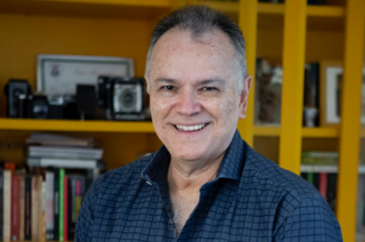 Imagem: Foto posada do diretor Wolney Oliveira, um homem branco utilizando uma camisa social azul e ao fundo uma estante com livros e câmeras fotográficas