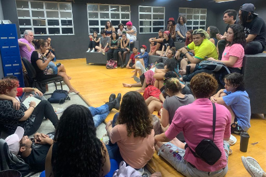 Imagem: Em uma sala, jovens estão sentados no chão formando um semi círculo enquanto duas pessoas estão sentadas em cadeiras fazendo algumas explanações