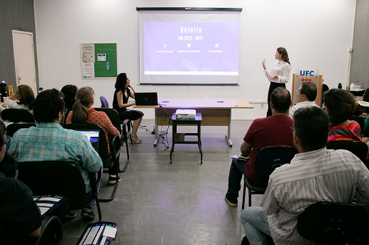 Imagem: Pessoas em um auditório; em pé, apresentando slides, está a servidora Roberta Queirós; ao lado dela está sentada a servidora Kamila Motta; em frente a elas estão os gestores, sentados em cadeiras (Foto: Ribamar Neto/UFC)