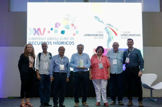 Imagem: sete pessoas estão em pé, em um palco. Elas vestem roupas sociais e usam crachás. Ao centro, um homem, de camisa azul, segura uma escultura