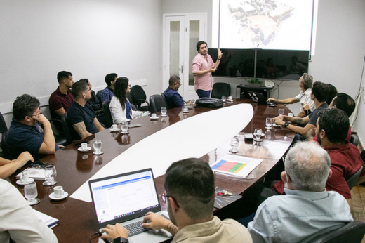 Imagem: Foto da apresentação do projeto arquitetônico da Estação Científica da UFC em Jericoacoara com uma tela projetada e os participantes da reunião sentados em uma grande mesa oval