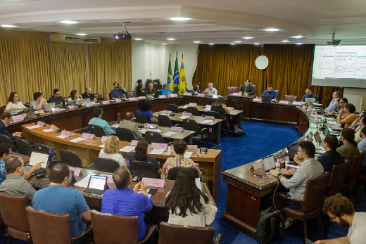 Imagem: foto de uma sala com carpete azul e mesas dispostas em círculo. Ao centro, três fileiras de mesas. Em todas as mesas há pessoas sentadas na cadeiras