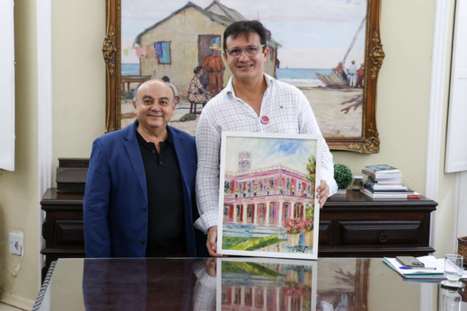 Imagem: Foto posada do artista Francisco Ivo vestido com um paletó azul marinho e o reitor Custódio Almeida de blusa branca e quadriculada de mangas comprida segurando um quadros 