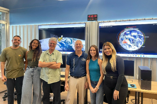 Imagem: estudantes e professores estão posicionados em pé, num laboratório com dois grandes monitores de computador atrás deles, que estão sorrindo para a foto.