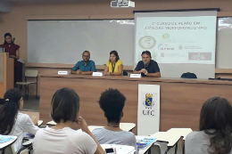 Imagem: foto de um auditório, de onde se vê pessoas sentadas de costas no público e, à frente delas, três pessoas na mesa do palco e uma à esquerda, em pé, atrás de um púlpito