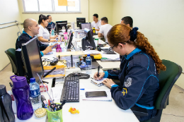 Imagem: foto de agentes da PM sentados em volta de uma mesa retangular, alguns usam computadores