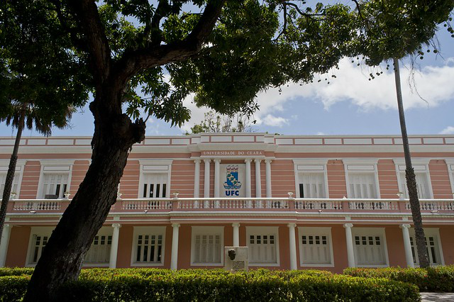 Imagem: Foto de vista lateral da Reitoria mostra os jardins e o brasão da UFC no andar superior; acima um céu azul com poucas nuvens e à frente, uma árvore. (Foto: Júnior Panela/ UFC Informa)