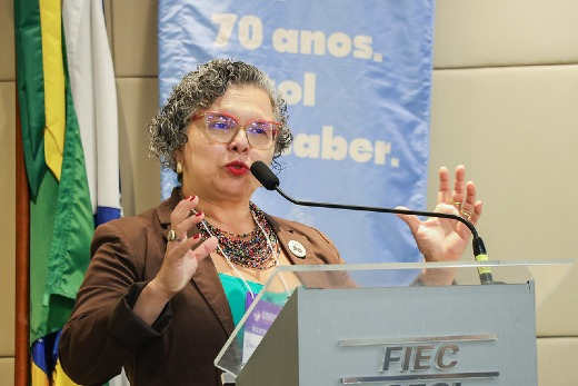 Imagem: Foto da professora Diana Azevedo, mulher branca, com cabelos grisalhos. Ela usa óculos vermelho e um blazer marron. Está gesticulando, enquanto fala em um púlpito onde se lê a palavra "Fiec"