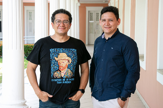 Imagem: Foto de dois professores autores dos artigos. Eles estão em pé, ambos com a mão no bolso da calça, em frente a um corredor com paredes de cor rosa e pilastras brancas (Foto: Divulgação)