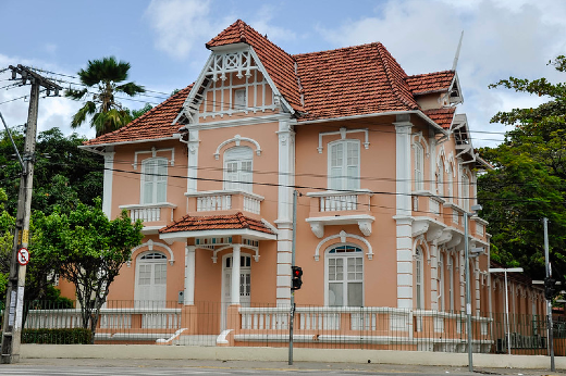 Imagem: Fotografia da Casa de Cultura Alemã, vista do outro lado da Avenida da Universidade; a casa é rosa, de arquitetura europeia e possui dois andares. (Foto: Ribamar Neto/ UFC Informa)