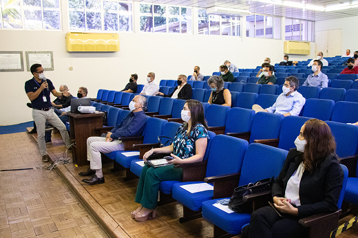 Imagem: foto de um homem de blusa polo azul faz apresentação em um auditório com pessoas sentadas de forma espaçada e com máscaras