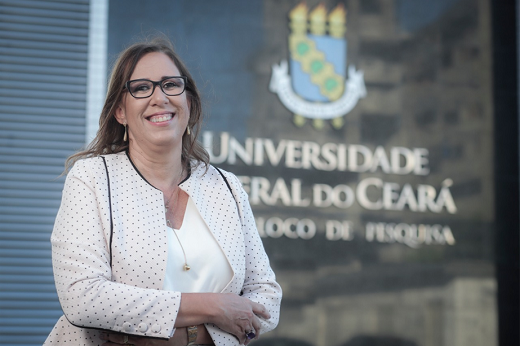 Imagem: Foto da professora Cláudia do Ó Pessoa. Ela veste uma roupa branca em frente a uma placa com a marca da UFC