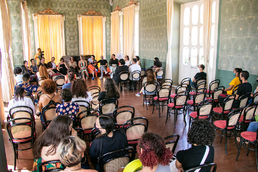 Imagem: Ambiente decorado com paredes verdes, janelas, cortinas, cadeiras de madeira e pessoas sentadas.