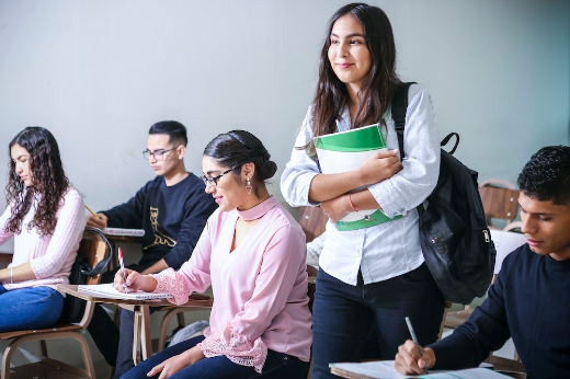 Imagem: aluna com livros na mão em pé, com outros estudantes em volta dela