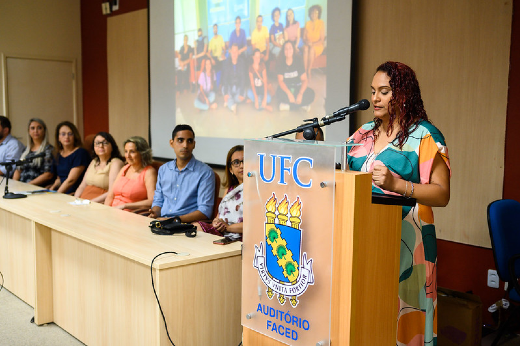 Imagem: Mulher com roupa colorida fala no microfone e ao lado mesa formada com outras pessoas sentadas e foto projetada ao fundo.