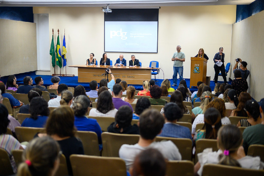 Imagem: auditório completo com plateia de costas e ao fundo mesa composta por algumas pessoas sentadas e outras de pé.