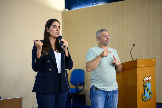 Imagem: mulher branca de cabelos lisos e escuros fala ao microfone e ao lado homem branco, cabelos grisalhos, com blusa branca faz a interpretação em libras.