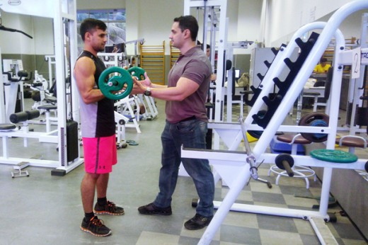 Imagem: dois homens em sala de musculação, um com roupas de ginástica segura um peso e outro acompanha