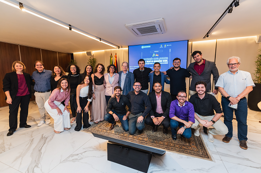 Imagem: Representantes da UFC, da UFSC e do Instituto Fraunhofer posam juntos, entre participantes em pé e agachados, em frente a um telão azul. (Foto: Laura Guerreiro/Ascom FIEC)