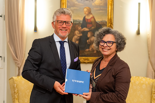 Imagem: De pé, no Salão dourado da Reitoria, o embaixador Andrii Melnyk e a vice-reitora Diana Azevedo sorriem e seguram juntos o livro Viva UFC, de capa azul. Ele veste terno escuro e ela, blazer marrom. (Foto: Viktor Braga/UFC Informa)