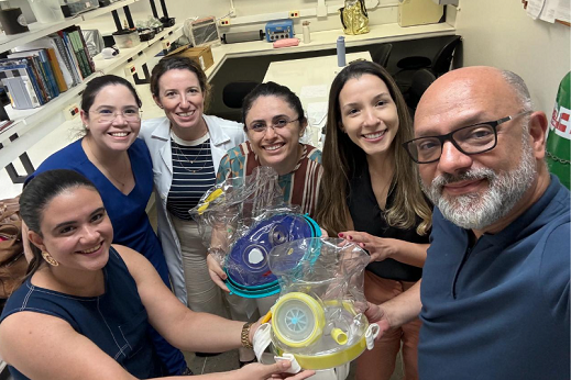 Imagem: O capacete Helmet Ped (amarelo), de origem italiana, foi inspiração para o Elmo (azul), criado no Ceará, que agora dão base para o Projeto Elminho. Na foto, o Prof. Marcelo Alcântara está com Luanna de Queiroz, médica intensivista pediátrica; Thaís Modesto, fisioterapeuta respiratória; Betina Tomaz, fisioterapeuta intensivista; Isabella Mattos, médica pneumologista; e Camila Leite, fisioterapeuta e docente da UFC, integrantes do RESPLAB (Foto: Marcelo Alcântara)