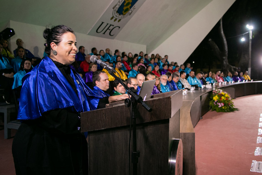 Imagem: mulher fala ao microfone com roupa azul e preta e mesa formada ao fundo