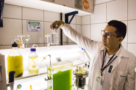 Imagem: Homem de jaleco segura, na altura do olhar, um frasco de vidro com líquido incolor, em frente a uma bancada de laboratório com várias peças de vidraria e equipamentos