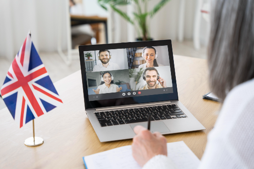 imagem: Mesa de trabalho na cor marrom com um notebook e uma bandeira da inglaterra e uma mulher sentada à frente com blusa branca e cabelos lisos.