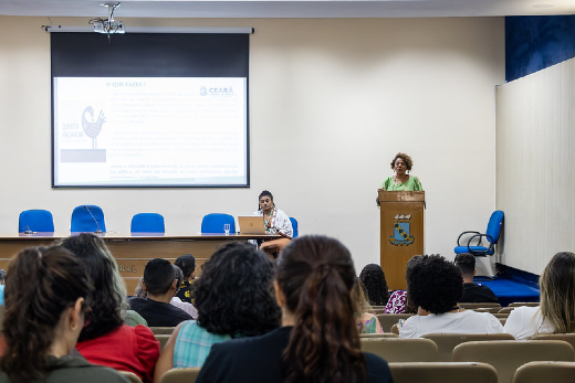 Imagem: A imagem mostra uma apresentação em um auditório. Uma mulher está em pé, falando em um púlpito à direita, enquanto outra pessoa está sentada à mesa à esquerda, com um laptop. Atrás delas, há uma tela de projeção com um slide exibindo texto e o logotipo do Governo do Ceará. O público está sentado, assistindo à apresentação. O ambiente tem cadeiras de cor clara e algumas bandeiras ao fundo. A pessoa no púlpito veste uma roupa verde-clara.