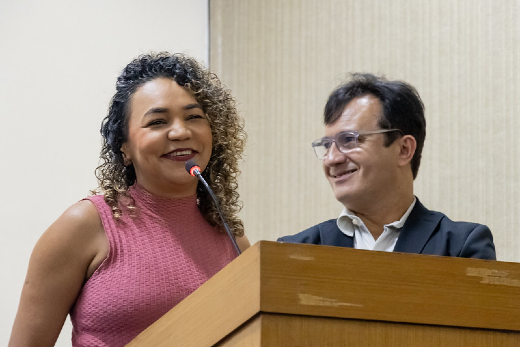 Imagem: A imagem mostra duas pessoas em pé atrás de um púlpito de madeira. A mulher à esquerda está sorrindo e falando ao microfone, vestindo uma blusa sem mangas rosa. O homem ao lado dela, à direita, também está sorrindo e usando óculos e um terno escuro com camisa branca. Eles parecem estar em um ambiente formal, durante discurso em um auditório. O fundo é composto por uma parede de cor neutra.