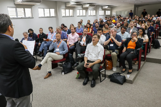 Imagem: A imagem mostra um grupo de pessoas sentadas em um auditório, assistindo a uma palestra ou apresentação. O orador está de costas, falando para a audiência, que parece estar atenta. As pessoas estão em assentos dispostos em fileiras, e muitas delas estão vestindo crachás, sugerindo que pode ser uma conferência, seminário ou evento profissional. O ambiente é iluminado e moderno, com janelas ao fundo e sistema de ar-condicionado visível no teto.