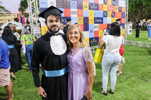 Imagem: homem de barba curta e bigode vestido com uma beca de formatura preta, acompanhada por uma faixa azul e um chapéu de graduação posa para foto sorrindo ao lado de sua mãe, uma mulher loira, que usa óculos e veste um vestido longo lilás, com uma parte de renda sobreposta em cima. Ao fundo, há um painel colorido com o logo da Universidade Federal do Ceará (UFC) e outros símbolos relacionados à formatura, junto com o texto "Formei!" e o brasão da universidade. A imagem parece ter sido tirada em um evento de formatura ao ar livre, sobre um gramado. 