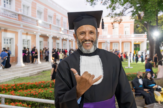 Imagem: homem negro, de meia idade, barba e cabelos grisalhos, posa para foto vestindo beca e capelo, sorrindo e fazendo o sinal de hang loose com uma das mãos. Ao fundo, aparece a fachada da Reitoria da UFC