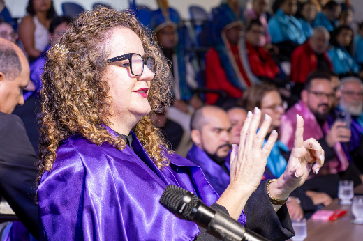 Imagem: mulher loira de cabelo cacheado e óculos, trajando veste talar na cor roxa, faz um sinal da Língua Brasileira de Sinais, ao lado de um púlpito