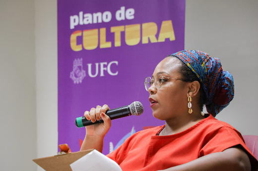 Imagem: Foto da palestrante Stephane Souto, mulher negra de vestido vermelho, sentada, com um microfone na mão. Ela está a frente com de banner roxo, onde se lê "Plano de Cultura da UFC", com letras brancas e amarelas (Foto: Ribamar Neto/UFC Informa)