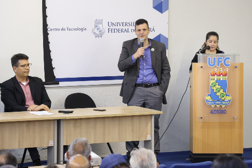 Imagem: homem de cabelos lisos e curtos, com calça e camisa sociais e blazer aparece falando ao microfone sobre um palco em um auditório, posicionado entre uma mesa comprida e um púlpito, atrás do qual aparece uma mulher, mestre de cerimônia. Atrás dele aparece um banner onde se lê Centro de Tecnologia e Universidade Federal do Ceará