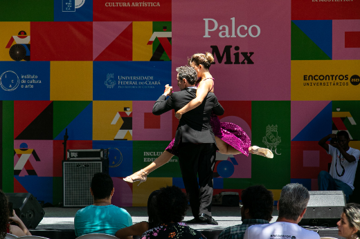 Apresentação de dança em um palco colorido ao ar livre, com a inscrição 'Palco Mix' ao fundo. Um casal de dançarinos está em movimento, com o homem, vestido de preto, segurando a mulher, que usa um vestido roxo brilhante e executa um movimento de salto com uma perna elevada. Na plateia, algumas pessoas assistem atentamente. Ao fundo, um fotógrafo registra a cena. O cenário do palco possui logos e padrões geométricos coloridos, representando um evento cultural.