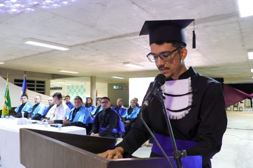 Imagem: homem jovem de pele parda vestindo beca está em púlpito discursando